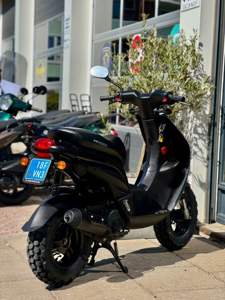 Zwarte Peugeot-scooter met grove terreinbanden geparkeerd voor een scooterwinkel in de zon.