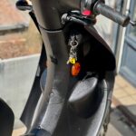 Close-up van een zwarte scooter met sleutels in het contact en oranje reflectoren, geparkeerd in de zon.