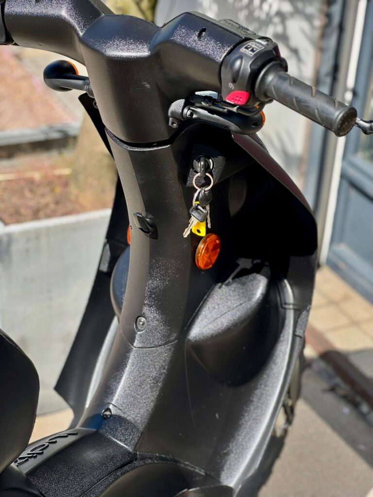 Close-up van een zwarte scooter met sleutels in het contact en oranje reflectoren, geparkeerd in de zon.
