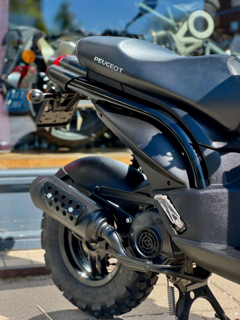 Close-up van de achterzijde van een zwarte Peugeot scooter met noppenband en uitlaat, geparkeerd bij een etalage.
