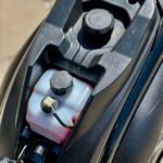 Close-up van scooter onder het zadel met brandstoftankdop en wit oliereservoir met slangetje.