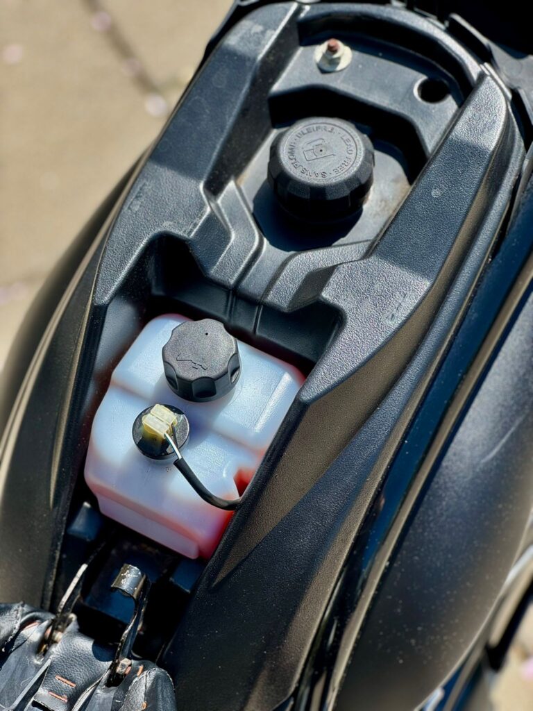 Close-up van scooter onder het zadel met brandstoftankdop en wit oliereservoir met slangetje.