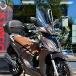 Bruine scooter met windscherm en topkoffer, geparkeerd voor een winkelruit op een zonnige dag.