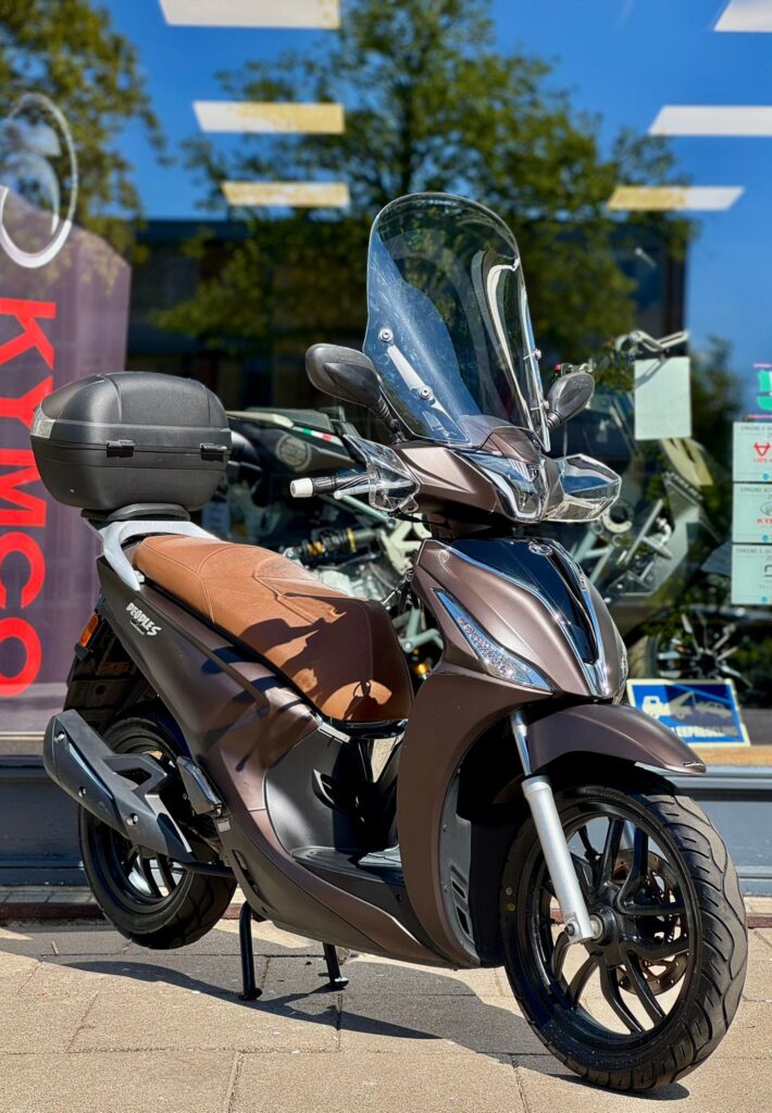 Bruine scooter met windscherm en topkoffer, geparkeerd voor een winkelruit op een zonnige dag.