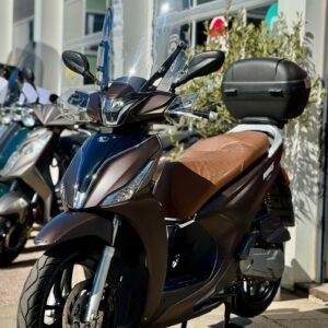 Donkerbruine scooter met hoog windscherm, bruin zadel en topkoffer, geparkeerd voor een showroom.
