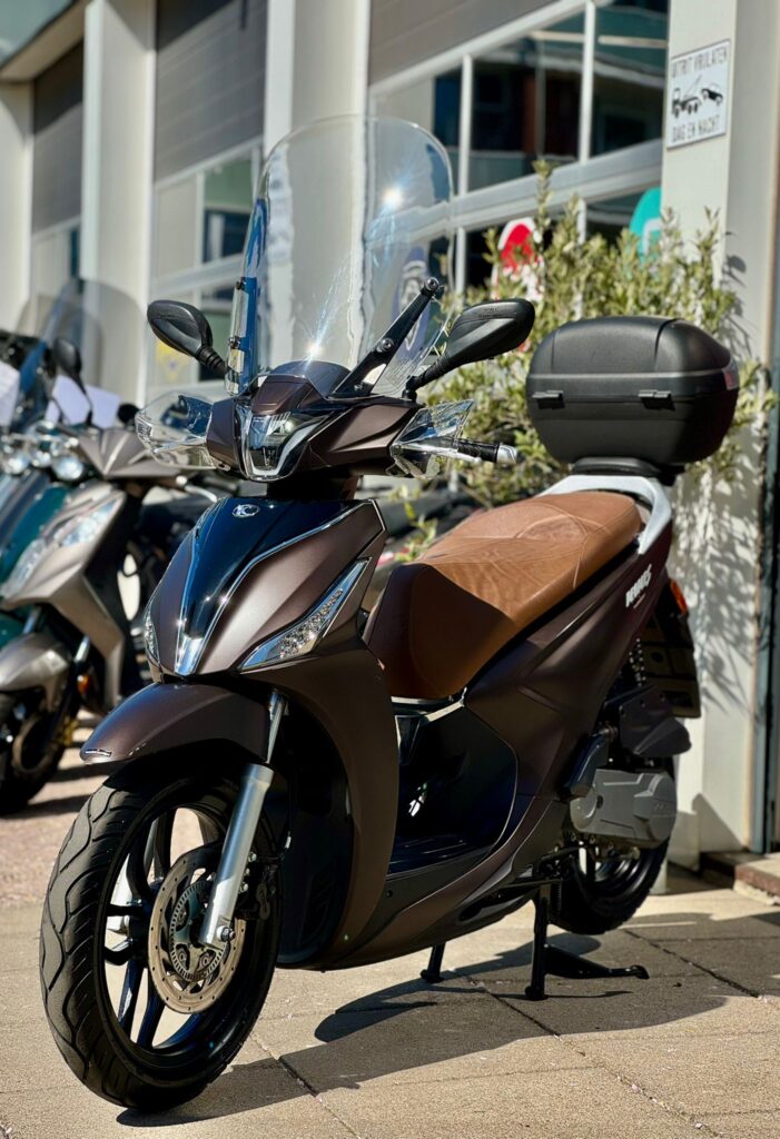 Donkerbruine scooter met hoog windscherm, bruin zadel en topkoffer, geparkeerd voor een showroom.