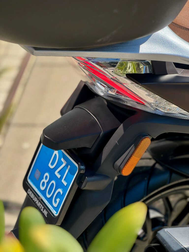 Close-up van het achterdeel van een scooter met achterlicht, oranje reflector en blauwe kentekenplaat.