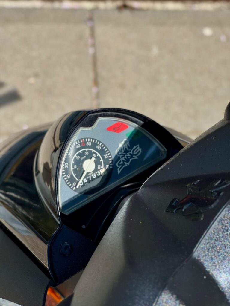 Close-up van het dashboard van een scooter met analoge snelheidsmeter en Peugeot-logo.