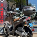 Bruine Kymco-scooter met topkoffer en ruit, geparkeerd op de standaard voor een showroom.