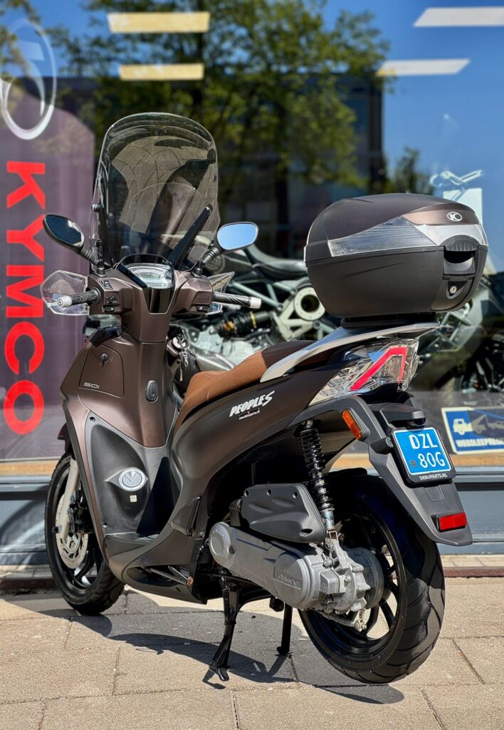 Bruine Kymco-scooter met topkoffer en ruit, geparkeerd op de standaard voor een showroom.