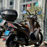 Bruine Kymco People S scooter met topkoffer geparkeerd voor een scootershop in de zon.