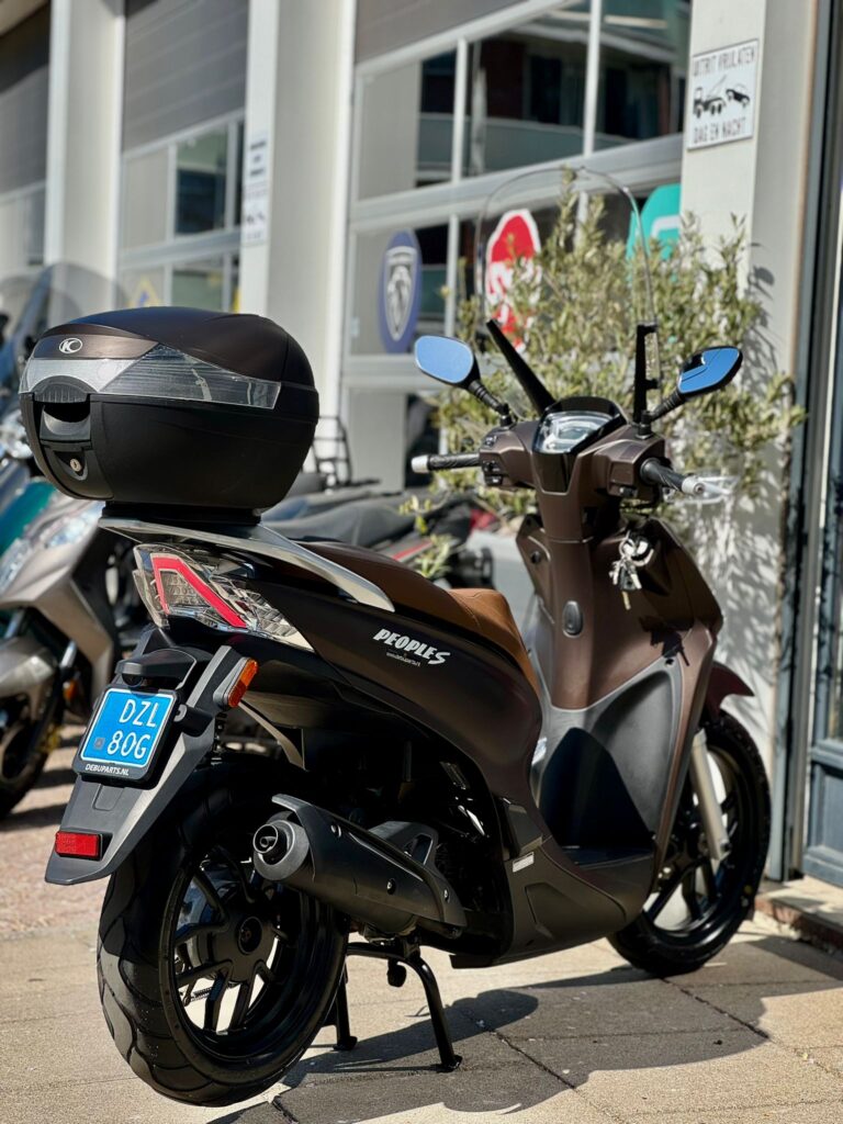 Bruine Kymco People S scooter met topkoffer geparkeerd voor een scootershop in de zon.