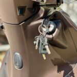 Close-up van een scooterslot met bungelende sleutels in het contact, op een matte bruine scooter.