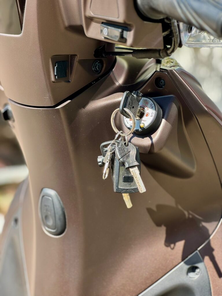 Close-up van een scooterslot met bungelende sleutels in het contact, op een matte bruine scooter.