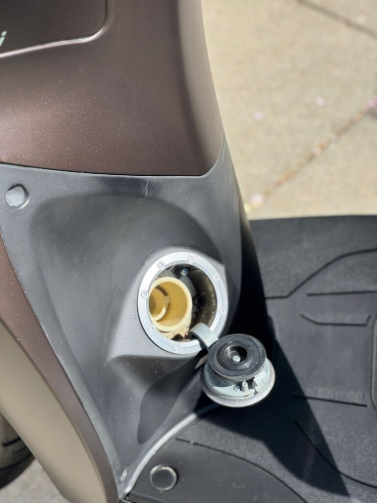 Open benzinedop van een scooter, vulopening zichtbaar naast de voetplaat.