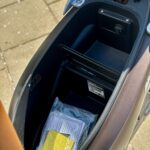 Open zadelbak van een scooter met opbergruimte en documenten in een plastic zak.