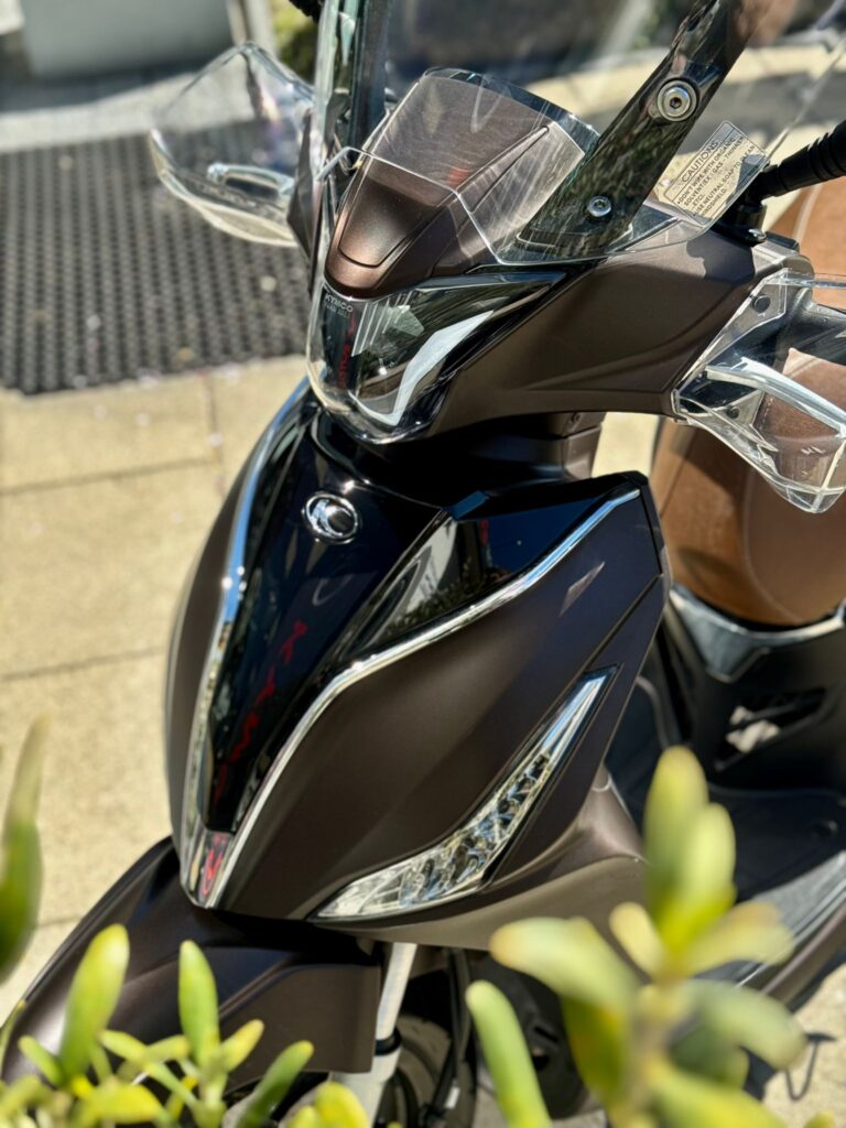 Close-up van de voorkant van een moderne matbruine scooter met windscherm en slanke LED-koplampen in de zon.