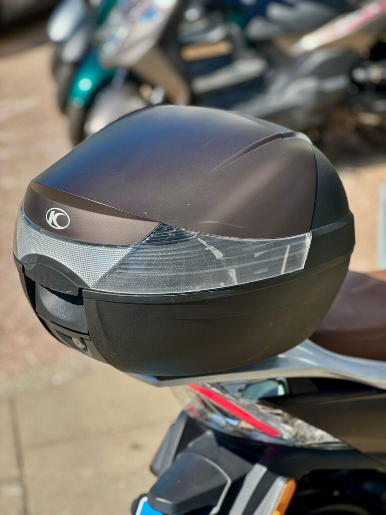 Close-up van een zwarte topkoffer op een scooter, met reflectoren en onscherpe scooters op de achtergrond.