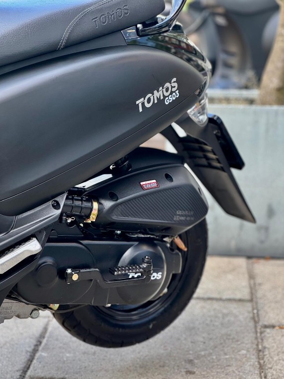 Close-up van de zijkant van een zwarte TOMOS GS03 scooter met zadel, uitlaat en achterwiel.