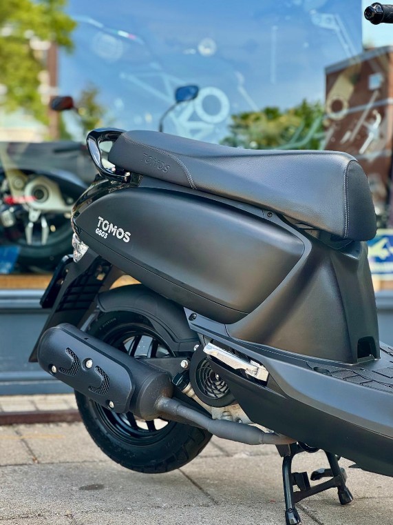 Matzwarte Tomos-scooter op de middenbok, geparkeerd voor een etalageraam.