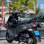 Zwarte scooter op standaard met blauw kenteken, geparkeerd voor een winkelruit in de stad.