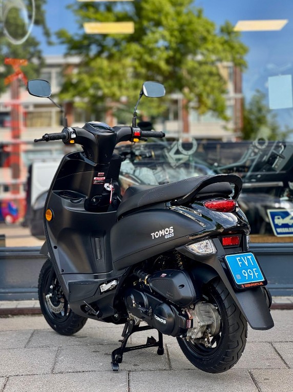 Zwarte scooter op standaard met blauw kenteken, geparkeerd voor een winkelruit in de stad.