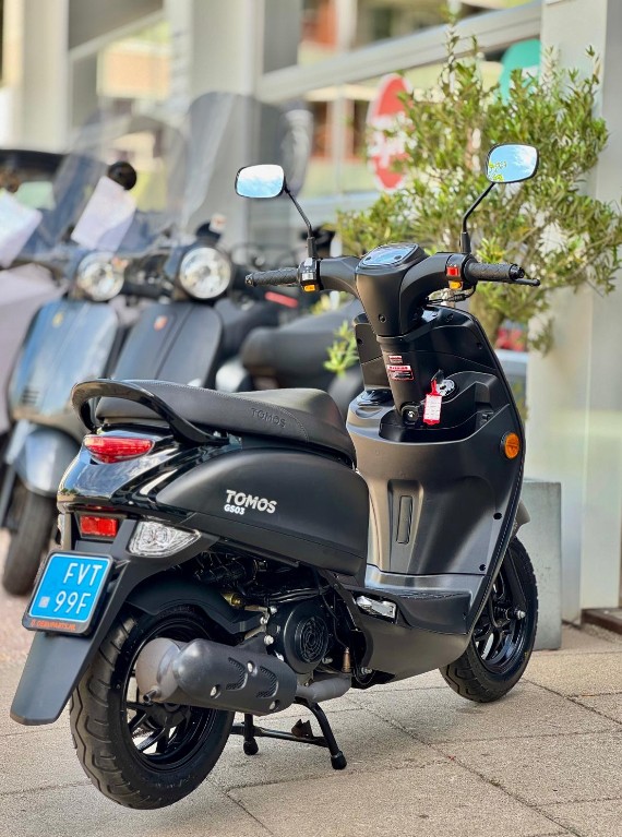 Zwarte Tomos 650S scooter met blauwe kentekenplaat, geparkeerd op de stoep bij een showroom.