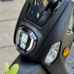 Close-up van de voorkant van een matte zwarte Tomos-scooter met koplamp en richtingaanwijzer.