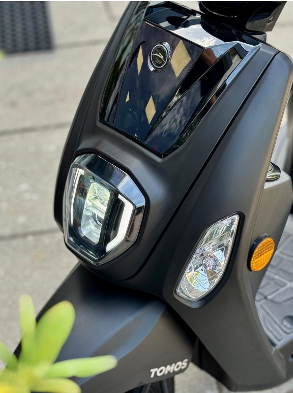 Close-up van de voorkant van een matte zwarte Tomos-scooter met koplamp en richtingaanwijzer.