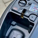 Opbergvak onder zadel van scooter, met tankdop en vergrendeling zichtbaar.