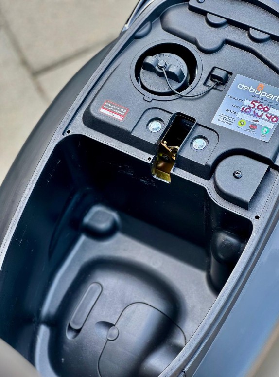 Opbergvak onder zadel van scooter, met tankdop en vergrendeling zichtbaar.