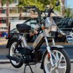 Klassieke grijze bromfiets met vierkante koplamp, geparkeerd op straat voor een winkelraam.