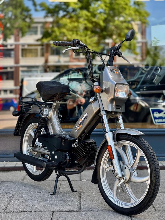 Klassieke grijze bromfiets met vierkante koplamp, geparkeerd op straat voor een winkelraam.