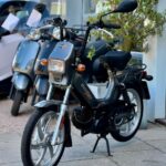 Zwarte retro brommer met rechthoekige koplamp, geparkeerd voor een rij scooters bij een winkel.