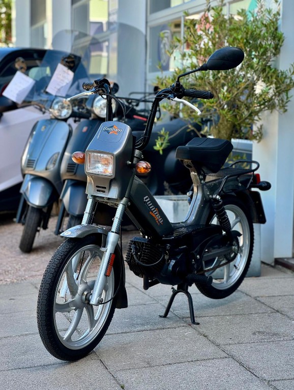 Zwarte retro brommer met rechthoekige koplamp, geparkeerd voor een rij scooters bij een winkel.
