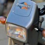Close-up van de koplamp met knipperlichten en Quadro by Tomos-logo op een grijze brommer.