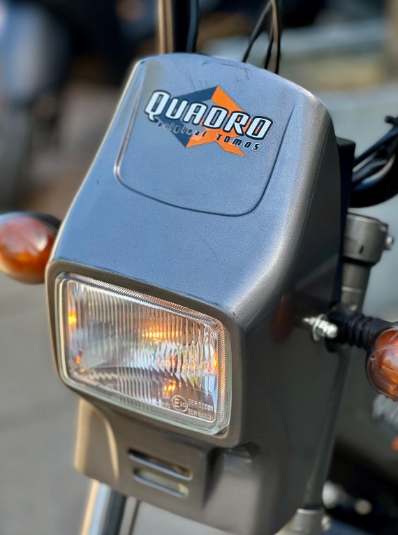 Close-up van de koplamp met knipperlichten en Quadro by Tomos-logo op een grijze brommer.