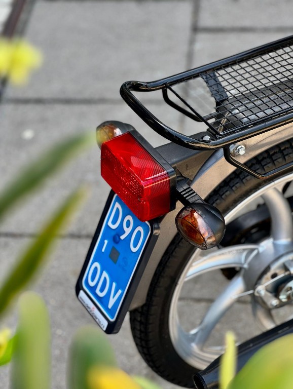 Close-up van de achterzijde van een bromfiets met bagagedrager, rood achterlicht en blauw kenteken.