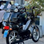 Klassieke grijze Tomos-brommer met blauwe kentekenplaat, geparkeerd voor scooters bij een showroom.
