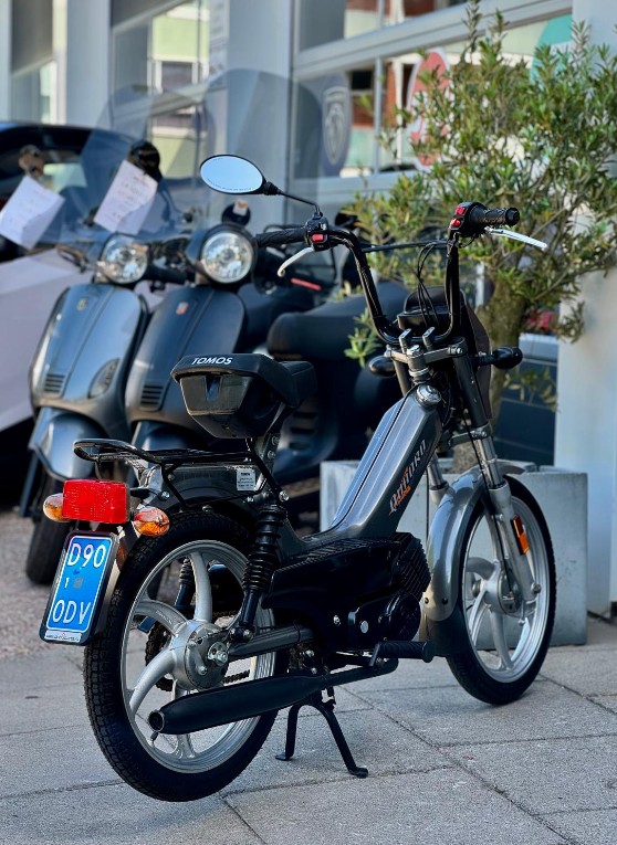 Klassieke grijze Tomos-brommer met blauwe kentekenplaat, geparkeerd voor scooters bij een showroom.