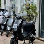 Zwarte scooter met hoog windscherm en bagagedrager, geparkeerd voor een showroom met andere scooters.