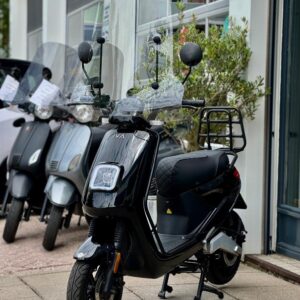 Zwarte scooter met hoog windscherm en bagagedrager, geparkeerd voor een showroom met andere scooters.