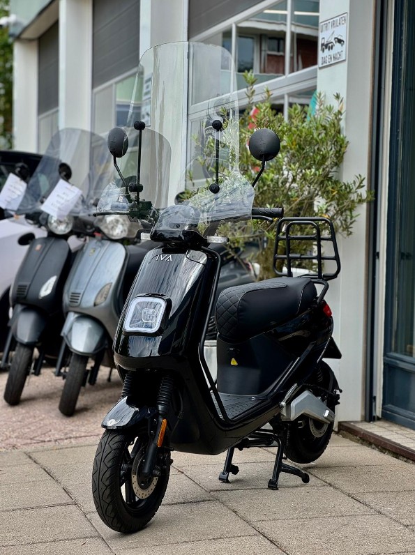 Zwarte scooter met hoog windscherm en bagagedrager, geparkeerd voor een showroom met andere scooters.