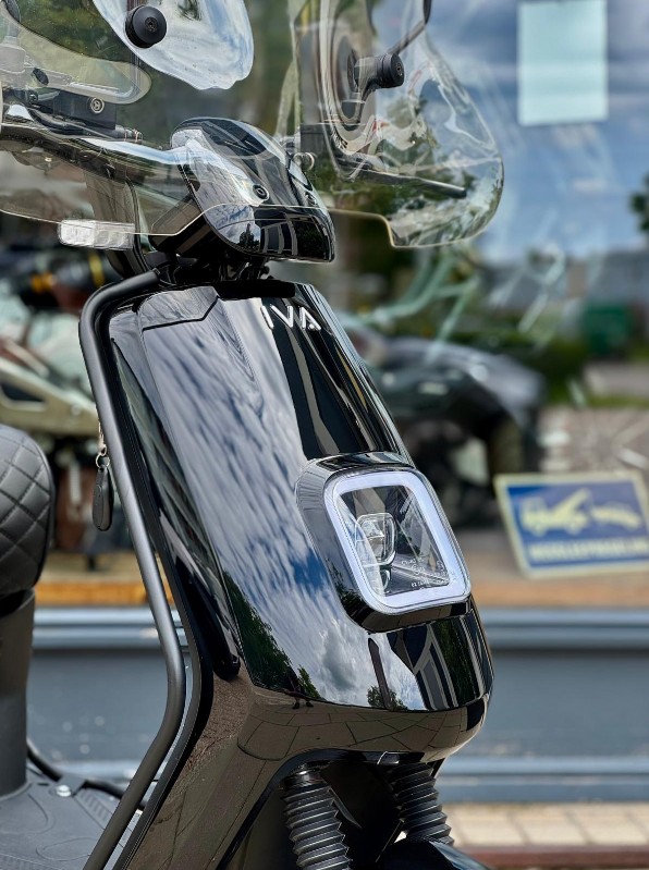 Close-up van de voorkant van een glanzend zwarte scooter met ruit en vierkante LED-koplamp.