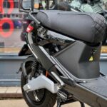 Close-up van zwarte elektrische scooter met bagagerek en gestikt zadel, geparkeerd voor een winkelruit.