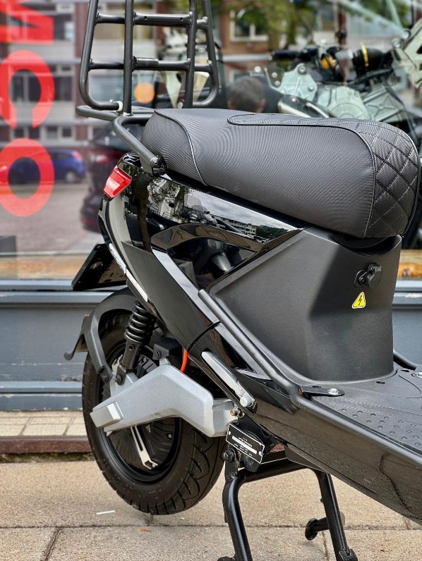 Close-up van zwarte elektrische scooter met bagagerek en gestikt zadel, geparkeerd voor een winkelruit.