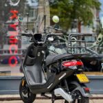 Zwarte Kymco-scooter met windscherm en bagagerek geparkeerd voor een etalage.