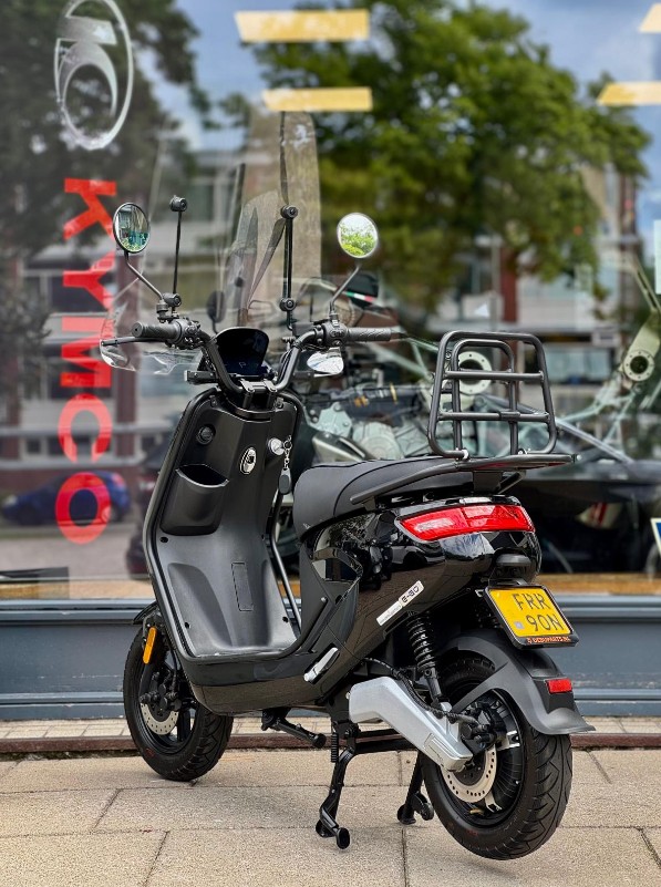 Zwarte Kymco-scooter met windscherm en bagagerek geparkeerd voor een etalage.
