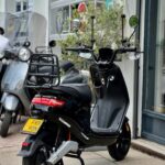 Zwarte elektrische scooter met bagagedrager en hoge spiegels, geparkeerd voor een winkel.