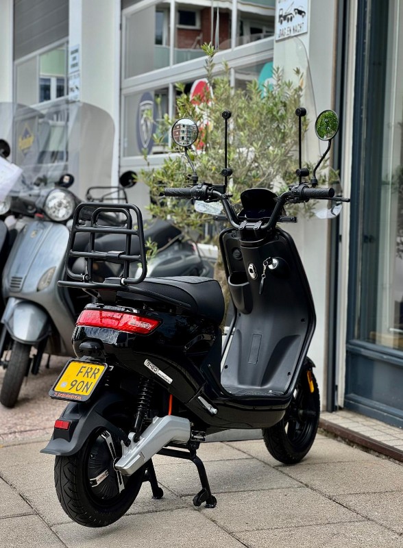 Zwarte elektrische scooter met bagagedrager en hoge spiegels, geparkeerd voor een winkel.